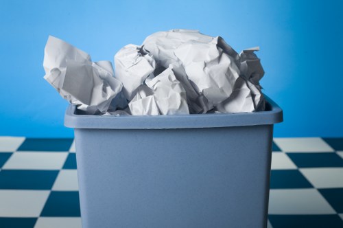 A rectangular light grey plastic bin filled with crumpled white paper, which appears to be used or discarded, situated on a black-and-white checkered floor inside a room or enclosed space. The background features a plain blue wall, providing a simple contrast to the bin and its contents. The paper is visibly wrinkled and loosely arranged, with some pieces leaning against the sides of the bin. The image captures the bin from a front-facing angle, highlighting the volume of waste material without showing any labels or additional objects. The overall setting suggests an indoor environment where rubbish collection or waste management services, such as those offered by Commercial Waste Morden, could be relevant for commercial or domestic waste removal purposes.