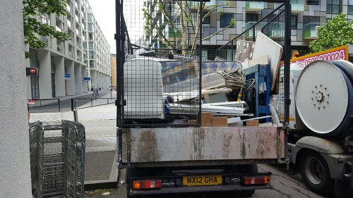 A flatbed truck with an open cargo area parked on a city street, loaded with assorted waste materials including large white panels, metal frames, plastic containers, and wooden planks, some of which are leaning against each other. The truck's rear is visible with a yellow registration plate reading 'NX12 GHA,' and the environment suggests an urban area with modern apartment or office buildings in the background. The surrounding pavement and a nearby sidewalk are partially visible, indicating a typical commercial waste removal scene likely serviced by Commercial Waste Morden as part of a rubbish removal operation in the Morden area.