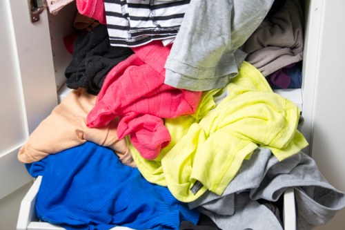 A close-up view of a partially open metal storage drawer filled with a variety of clothing items, including a bright yellow garment, a grey piece, and a blue piece, all made of different fabrics and textures. The clothes are disorganized and spilling over the edges of the drawer, which is situated on a white or light-colored surface, possibly within a storage unit or wardrobe. The setting suggests an indoor environment used for organizing personal items, unrelated to rubbish or waste removal. This image could be relevant to waste management services focused on collecting or disposing of clutter and clothing waste in a domestic or commercial setting near Morden, south London.