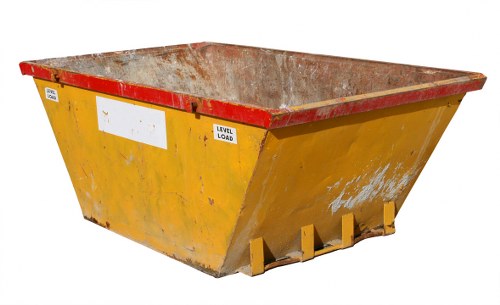 A large, industrial yellow skip with a red reinforced top edge, positioned on a paved surface, likely in a commercial or industrial area in Morden. The skip's exterior surface shows signs of wear with patches of chipped paint and dirt, indicating regular use for waste collection. The interior compartment is visible from the angle, revealing a rusty, metal-lined cavity designed for holding bulky rubbish. The bottom of the skip has four yellow support feet, slightly visible beneath the structure. The background appears neutral and uncluttered, emphasizing the skip as the main object. This image is relevant to rubbish removal services such as those provided by Commercial Waste Morden, highlighting typical equipment used for waste collection and disposal in a business or construction setting in the local area.
