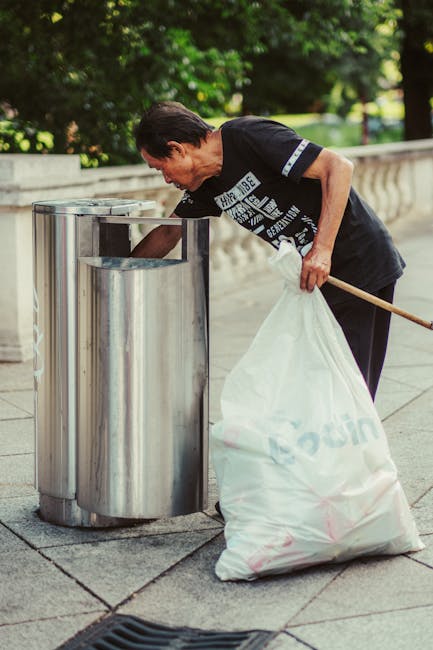 A middle-aged man with short dark hair, wearing a black T-shirt with graphic text and dark trousers, is seen leaning over a public stainless steel rubbish bin on a paved pathway. He is holding a large